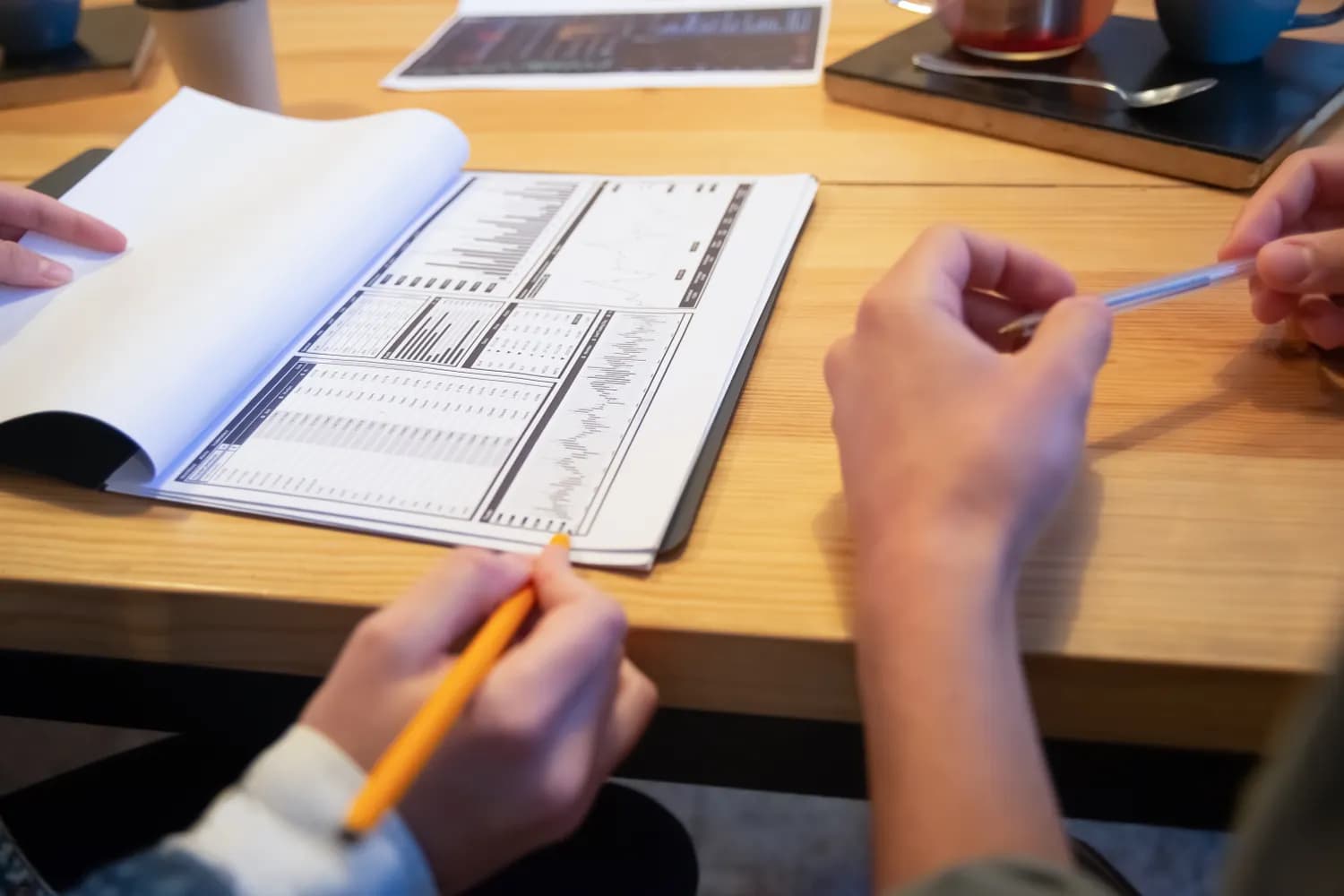 Deux personnes annotent au crayon un rapport imprimé de tableaux et de courbes, ouvert sur la table en bois d'un café